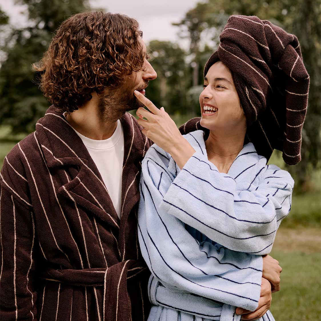 Two people in bathrobes and towels outdoors, one with a towel wrapped around their head.