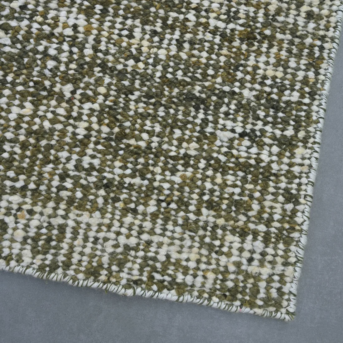 A close-up of a woven rug or textile showing a mottled pattern of olive green, cream, and white fibers, with a frayed edge, laid on a gray surface.