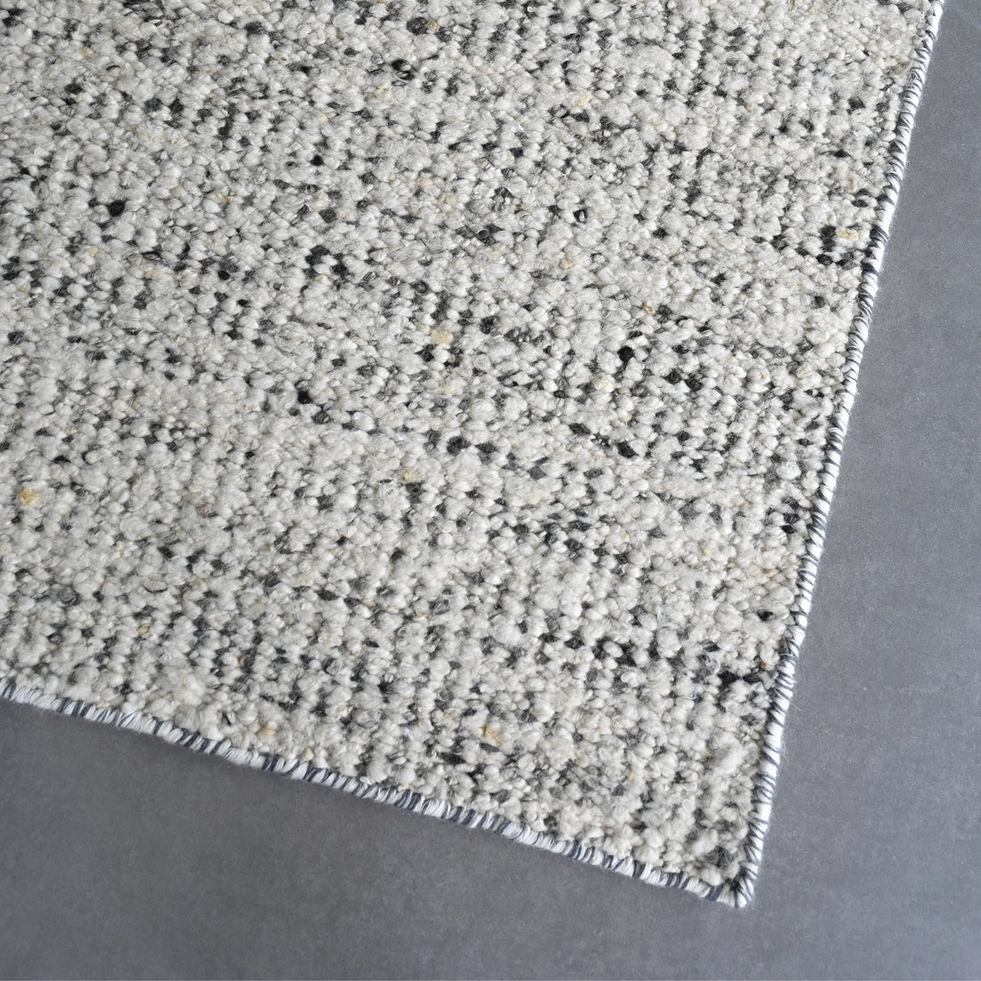 Close-up of a textured area rug corner showing a woven pattern in off-white, beige, and black with a fringed edge, placed on a gray floor.
