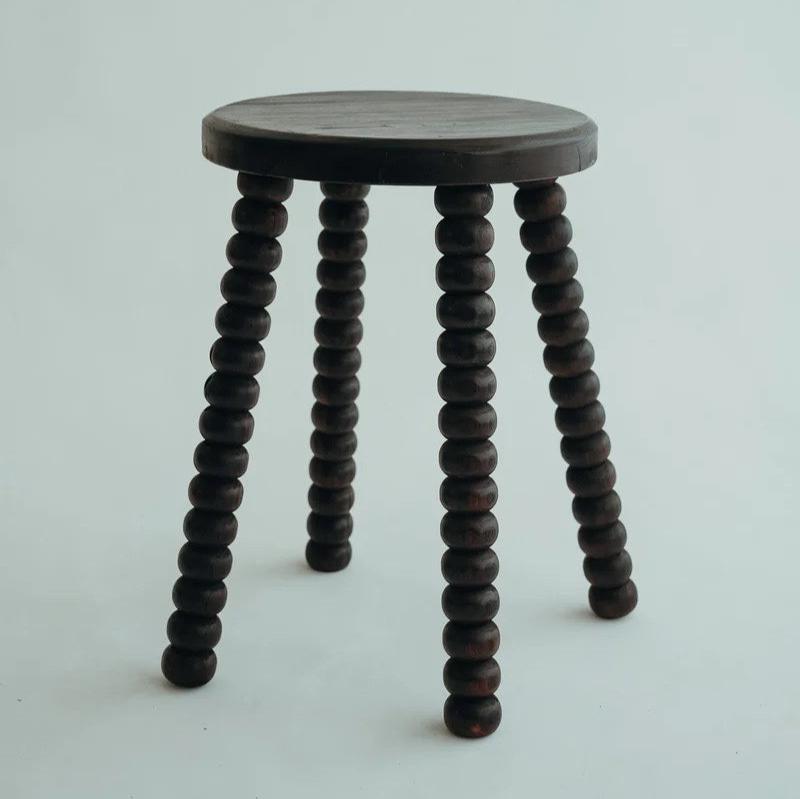 Carmen Stool in Walnut suar wood, round top on beaded bobbin-style legs, shown on a plain backdrop.
