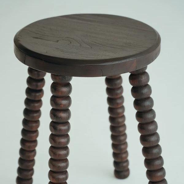Carmen Stool in Walnut suar wood, round top on beaded bobbin-style legs, shown on a plain backdrop.