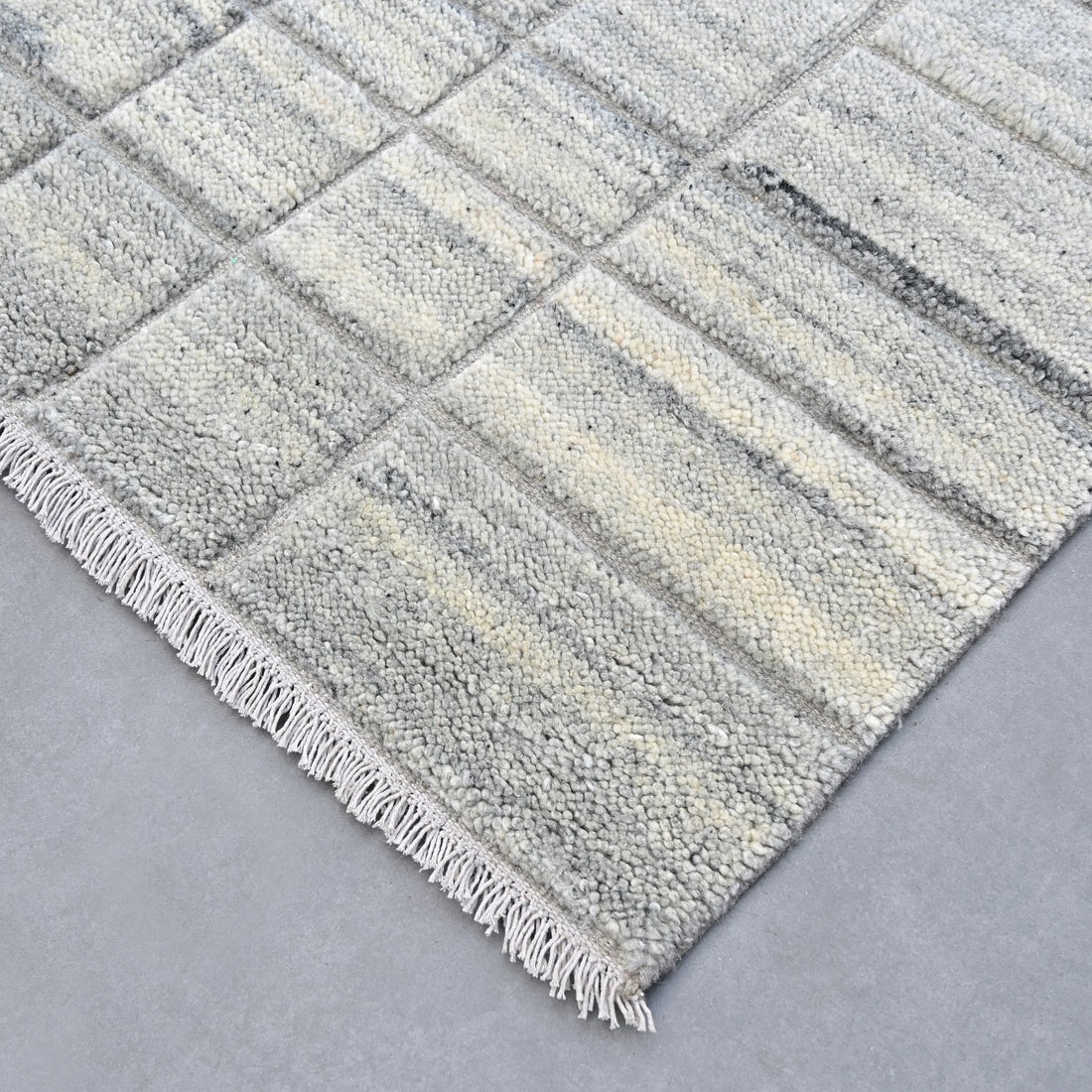 A close-up of a textured area rug with a woven, chunky striped pattern in shades of gray, beige, and off-white, finished with a fringed edge.