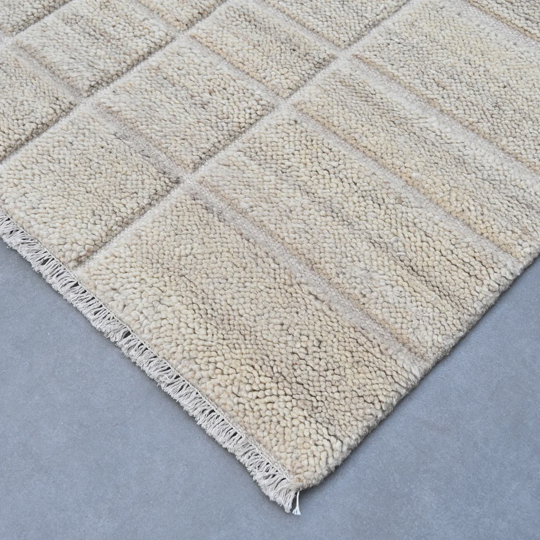 A close-up of a textured area rug with a chunky woven pattern and fringed edge, shown on a neutral gray floor.