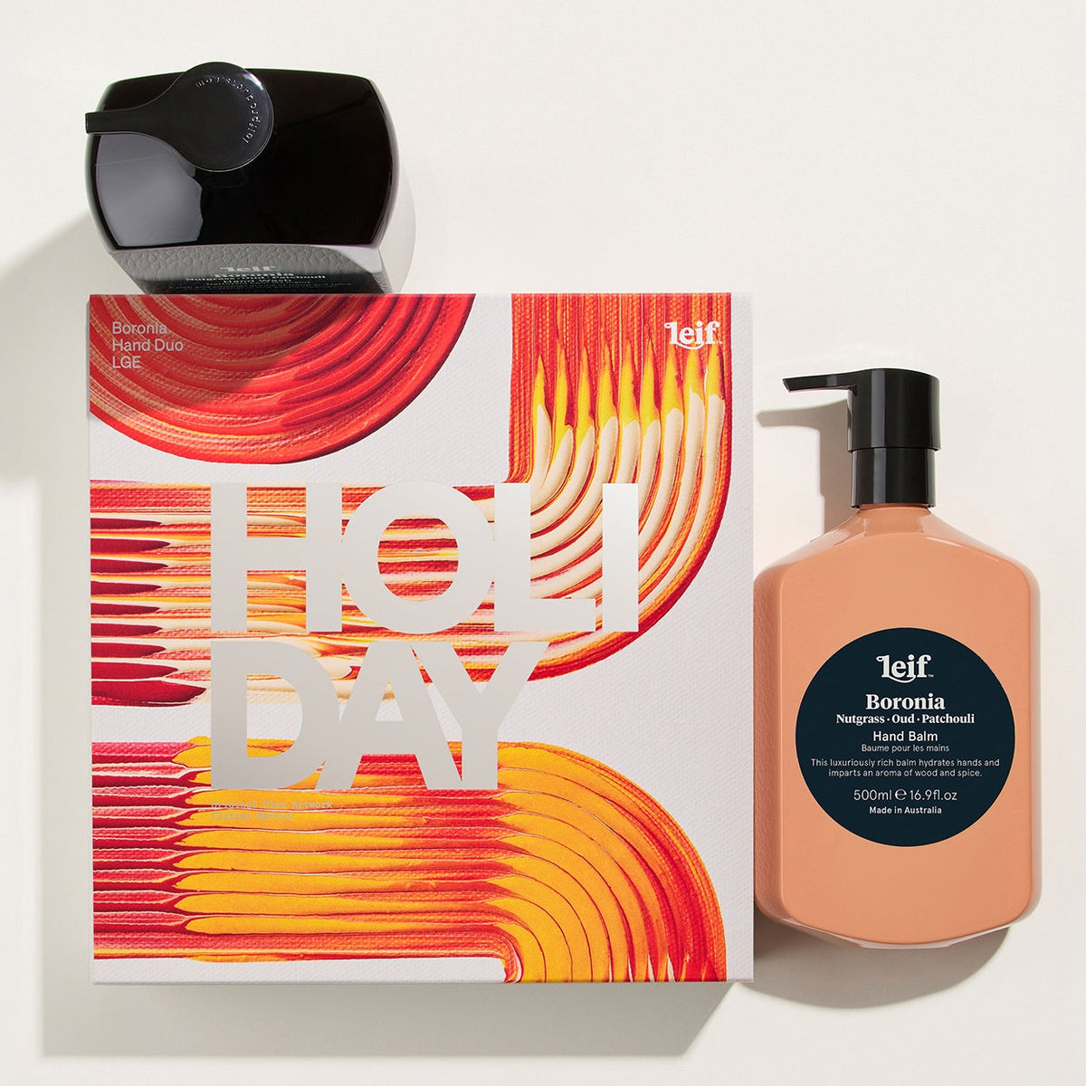 A 1elf holiday gift set featuring a black bottle of holiday light scented soy wax melts, a peach-colored bottle of Berrinia hand wash, and a white box with a vibrant red and orange abstract design and 'HOLIDAY' text, arranged on a clean white background.