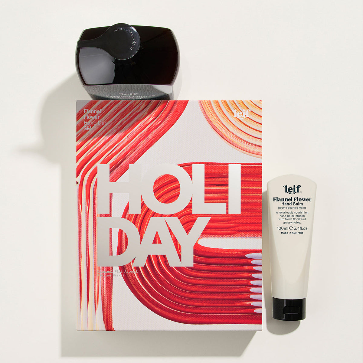 Leif Flannel Flower Hand Cream and matching gift set on a white background, featuring a white box with bold red and orange abstract lines and the word 'HOLIDAY' in large white letters, a black hand cream tube, and a black cap.