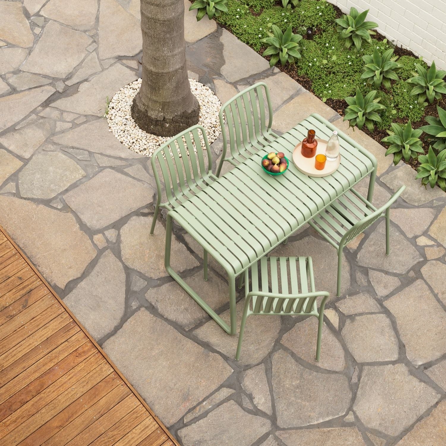 A green outdoor table with four chairs set on a stone-paved area with a tree in the background and some fruits on the table.