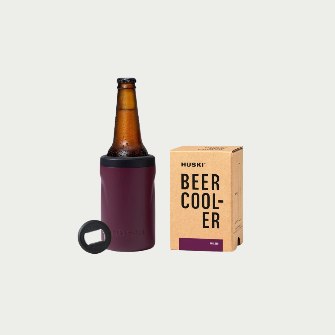 A Huski insulated beer cooler in deep plum purple shown with an amber glass beer bottle inside and its kraft paper packaging box on a light gray background.