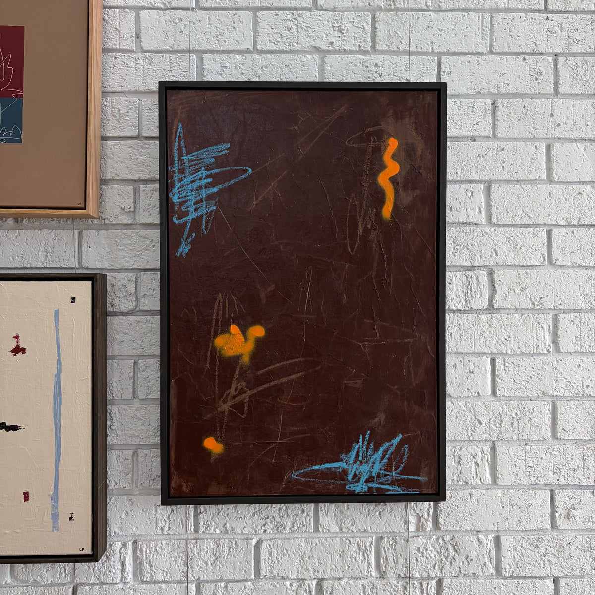 An abstract framed painting with a dark brown background and blue, orange, and yellow scribbled marks, hanging on a white brick wall next to other framed art.