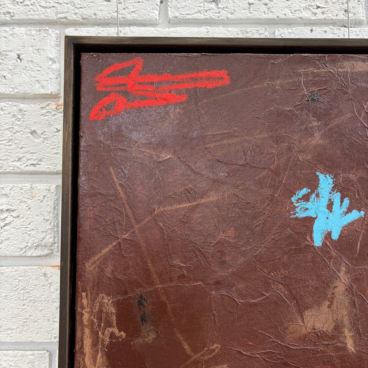 A square abstract painting with a textured dark brown background, featuring red and light blue scribbled marks, mounted on a white brick wall.