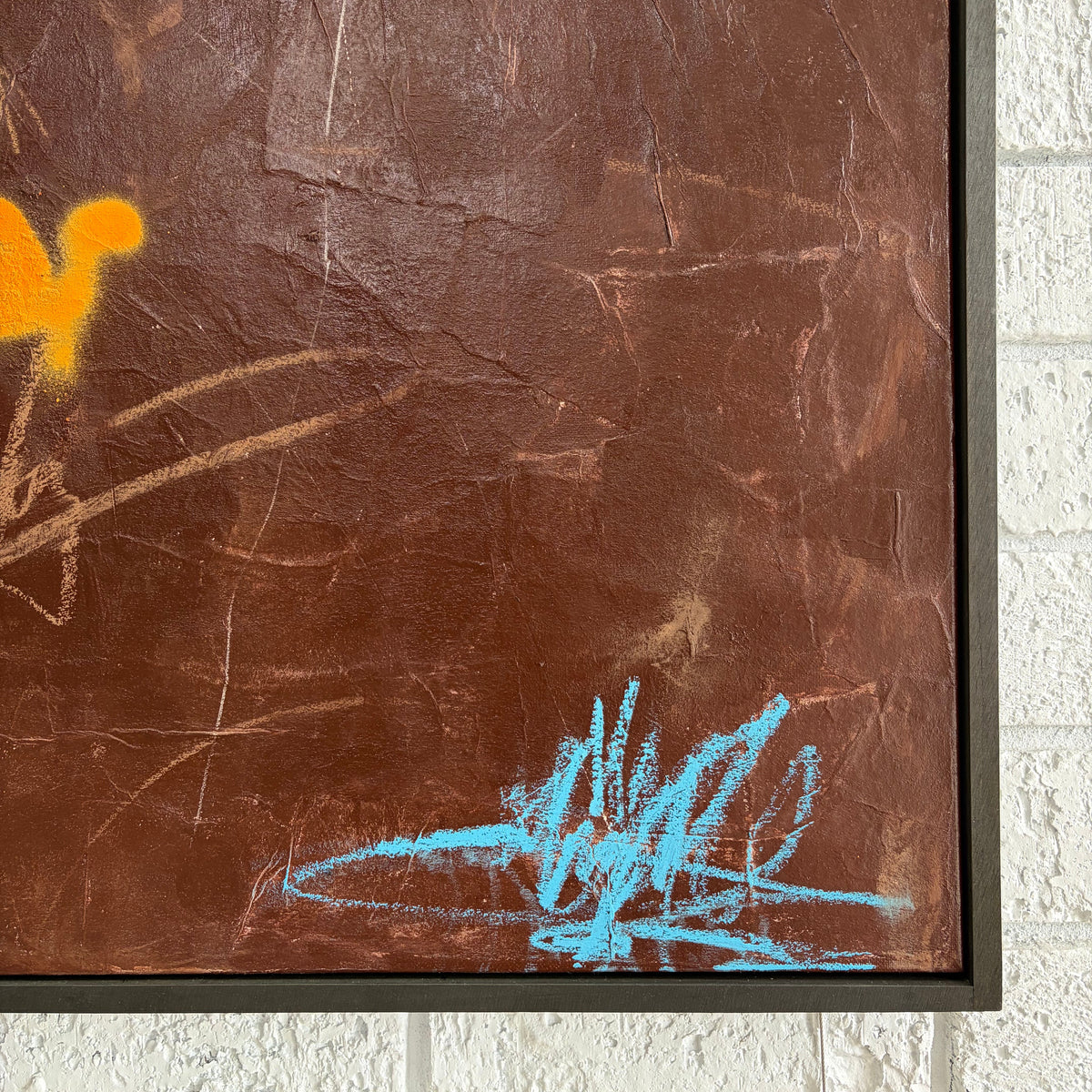 A close-up of a framed abstract artwork featuring a textured brown background with orange and light blue scribbled marks, mounted against a white brick wall.