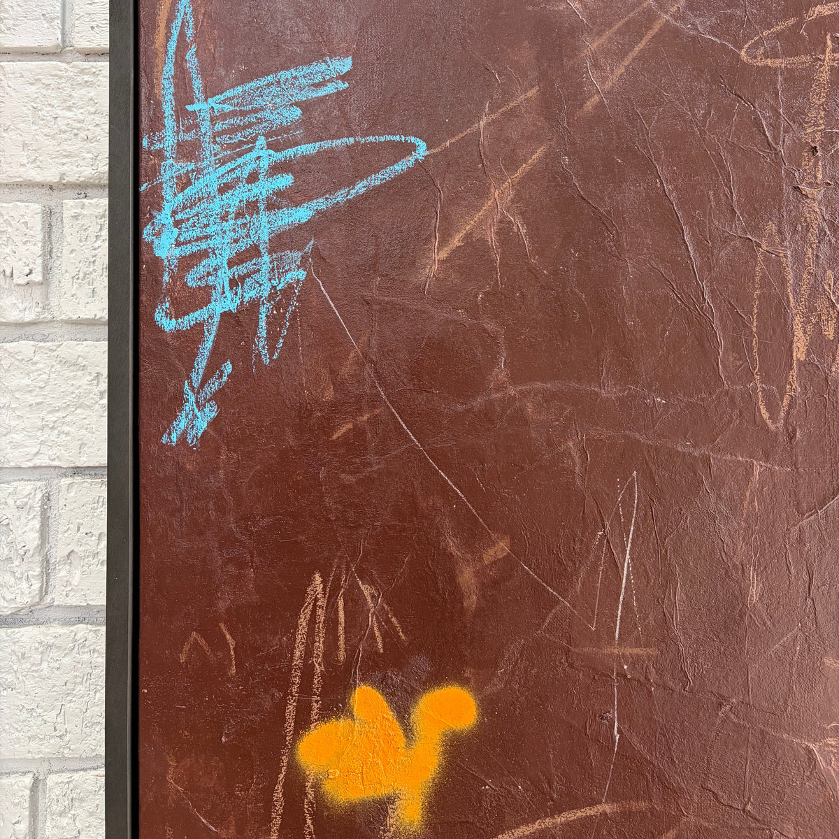 A framed abstract artwork featuring a textured brown background with light blue and orange scribbled marks, set against a white brick wall.