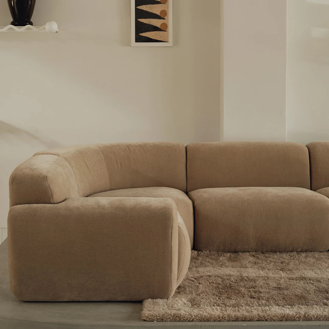 A modern curved modular sofa upholstered in a warm taupe-brown bouclé fabric, set in a minimalist living room with a shag rug and neutral walls.