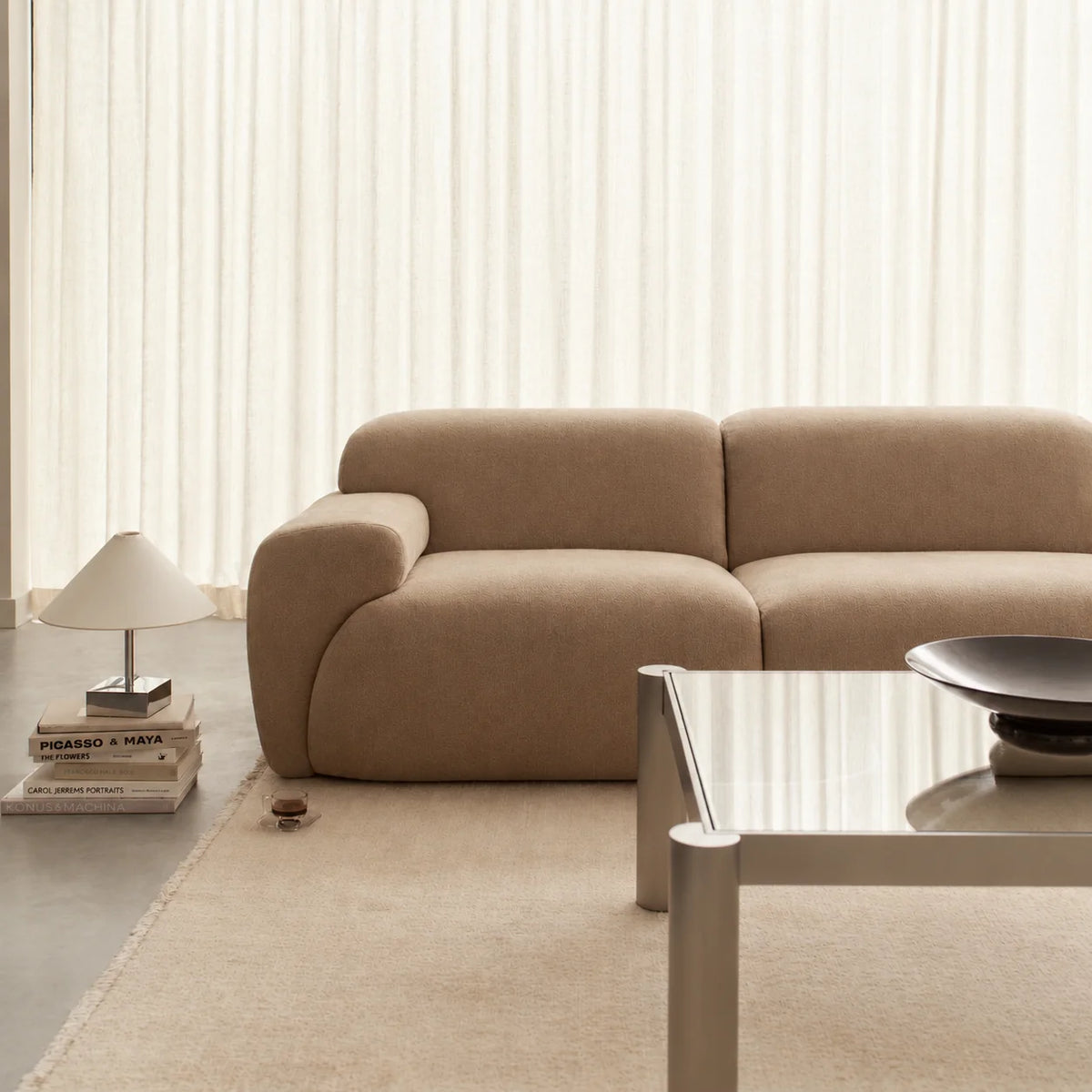 A modern beige upholstered sofa in a minimalist living room setting, positioned next to a glass coffee table and a small table lamp on a light rug.