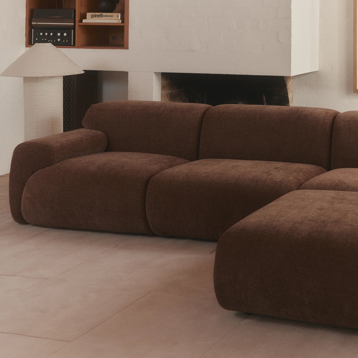 A modern modular sectional sofa in a warm brown bouclé fabric, styled in a minimalist living room with a white brick fireplace and neutral tile flooring.