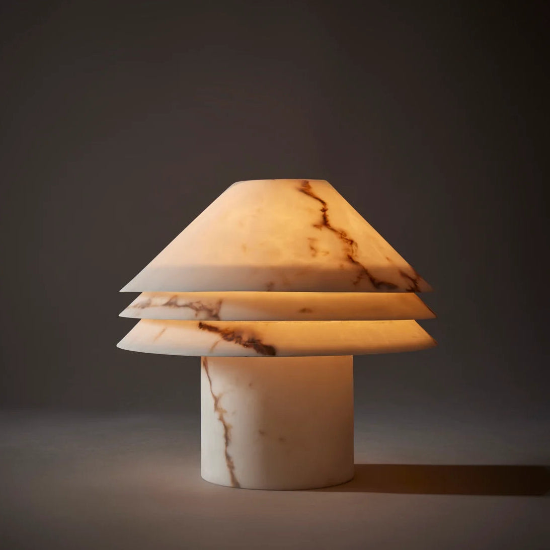 A modern table lamp made from polished onyx or marble, featuring a tiered conical shade and a cylindrical base, glowing with warm light against a dark gray background.