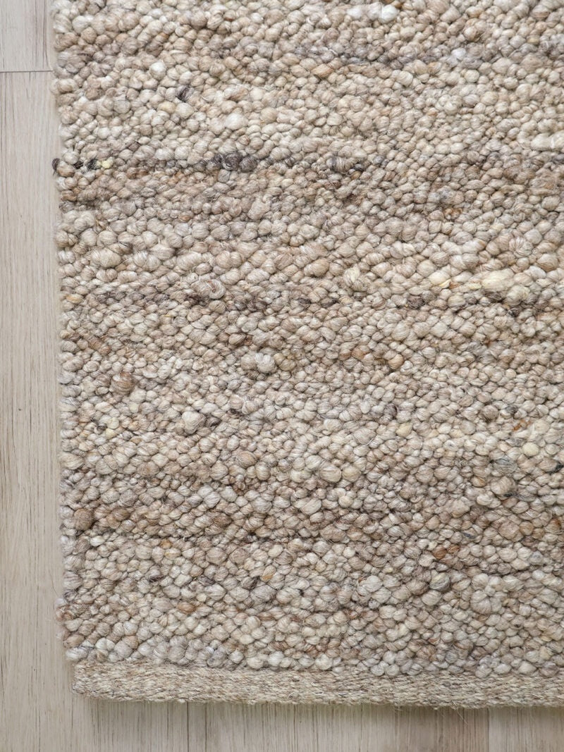 A close-up of a textured area rug with a chunky, pebble-like woven pattern in natural beige, cream, and light brown tones, shown on a light wood floor.