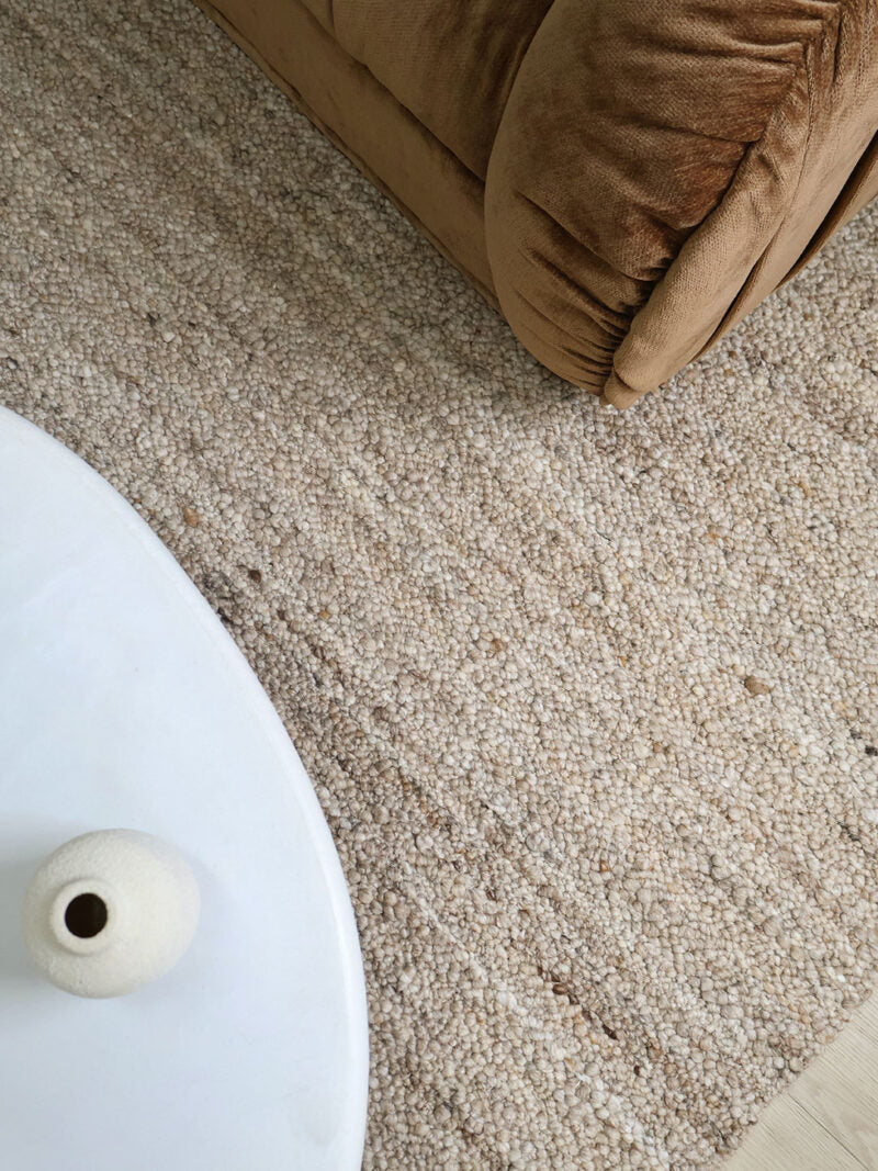 A close-up, high-angle view of a textured beige and brown woven area rug, showing its detailed loop pile. The bottom-left corner features a white curved table edge with a small white vase, and the top-right corner shows the corner of a brown velvet sofa.