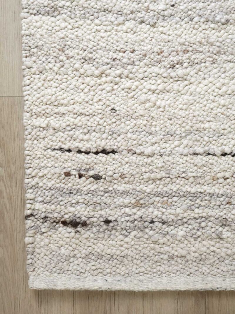 A close-up of a textured off-white and cream woven rug with subtle horizontal dark brown accents, shown on a light wood floor.