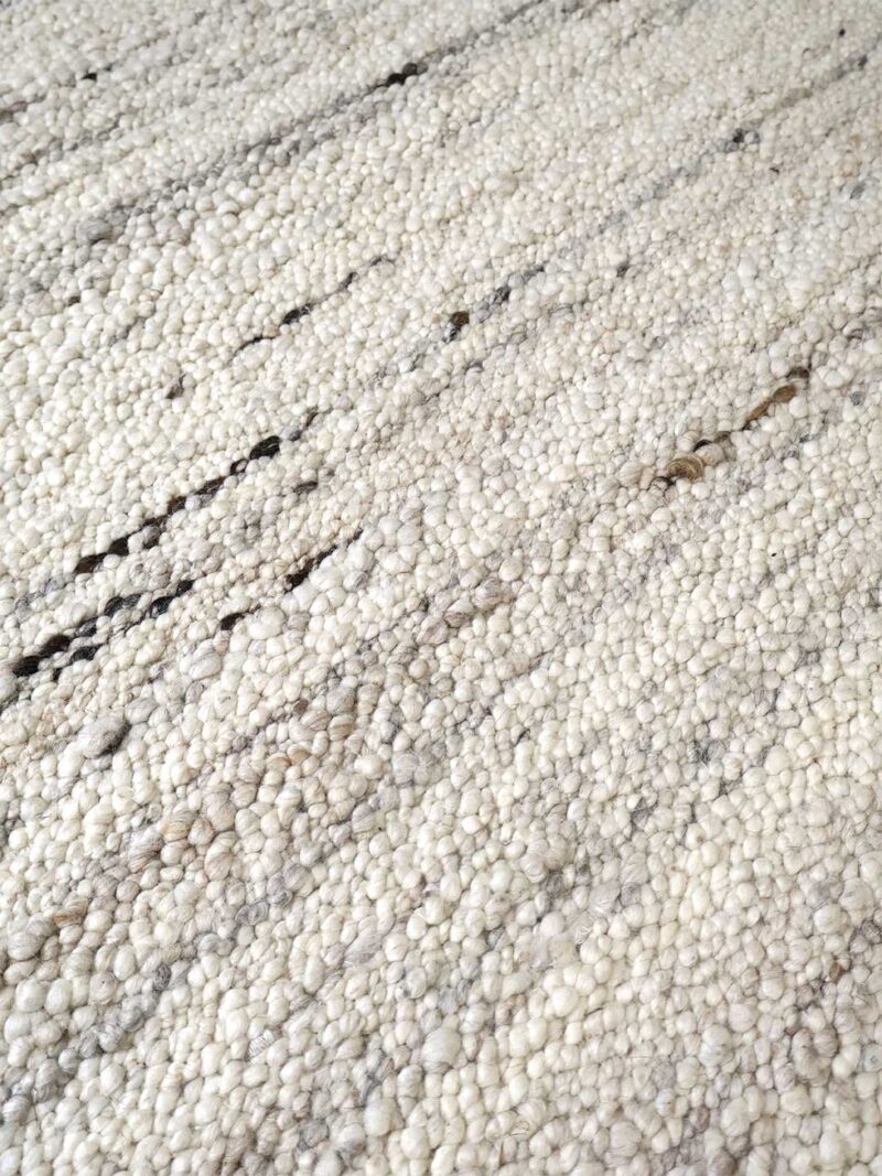 Close-up of a textured area rug with a dense, pebble-like pile in off-white, cream, and light gray with subtle dark brown and black streaks.