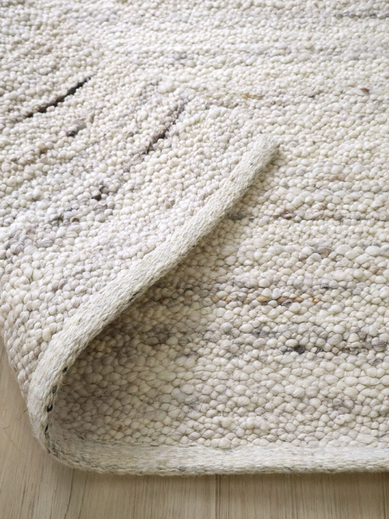 A close-up of a textured off-white woven rug with a curled corner, showing its chunky boucle-like weave and subtle gray flecks, placed on a light wood floor.