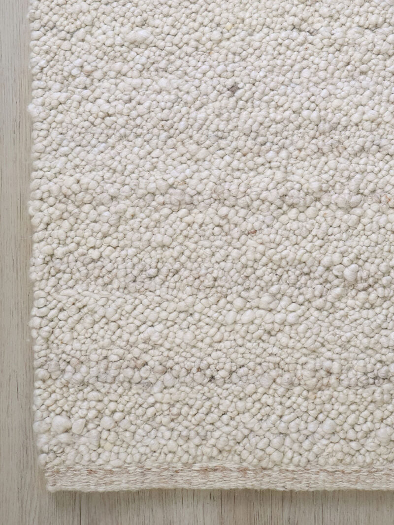 A close-up of a textured, chunky loop-pile rug in an off-white or cream color, laid flat on a light-colored wooden floor.