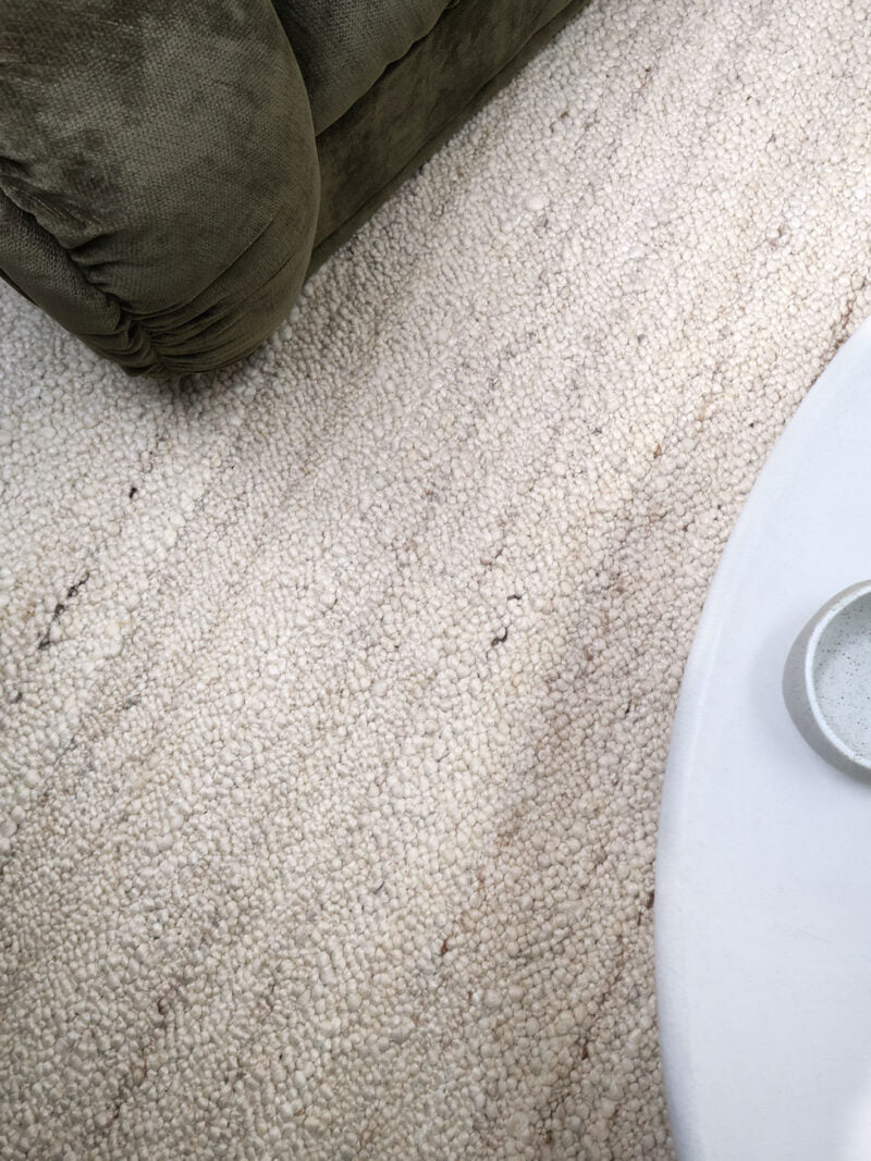 A close-up view of a textured, loop-pile area rug in a warm off-white or cream color, shown in a room setting with parts of a dark olive green upholstered piece of furniture and a white table visible.