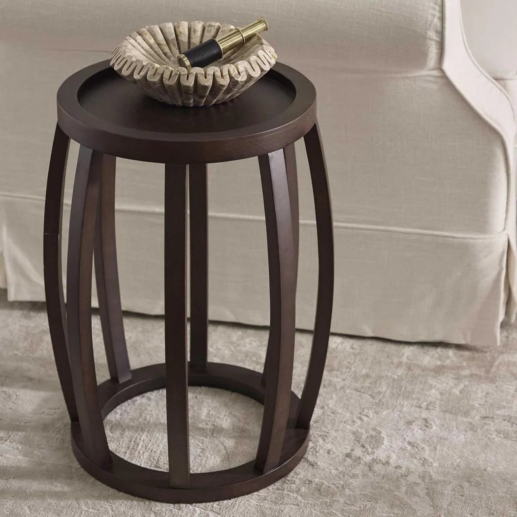 A dark brown wooden barrel-style side table with a round tray top, shown in a living room setting next to a beige sofa with a decorative bowl on top.