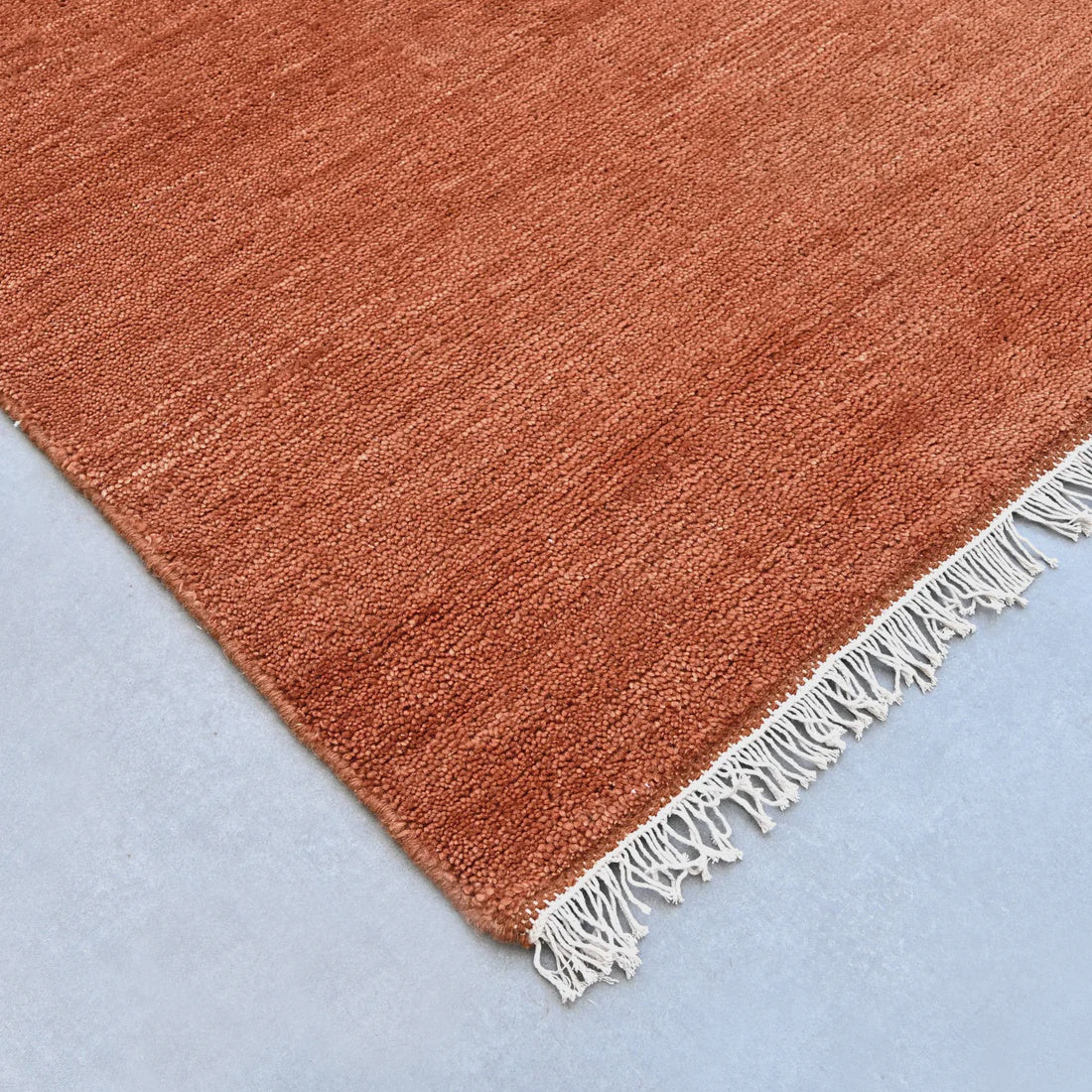 A close-up of a terracotta orange woven rug corner with white fringe, laid on a light blue surface.