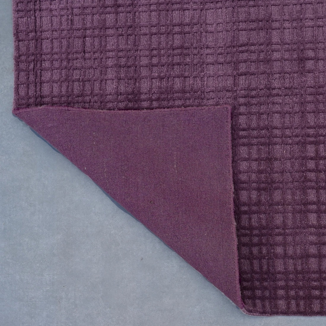 A close-up of a textured purple fabric, likely a rug or mat, with one corner folded over to show the solid purple backing, set against a neutral gray background.