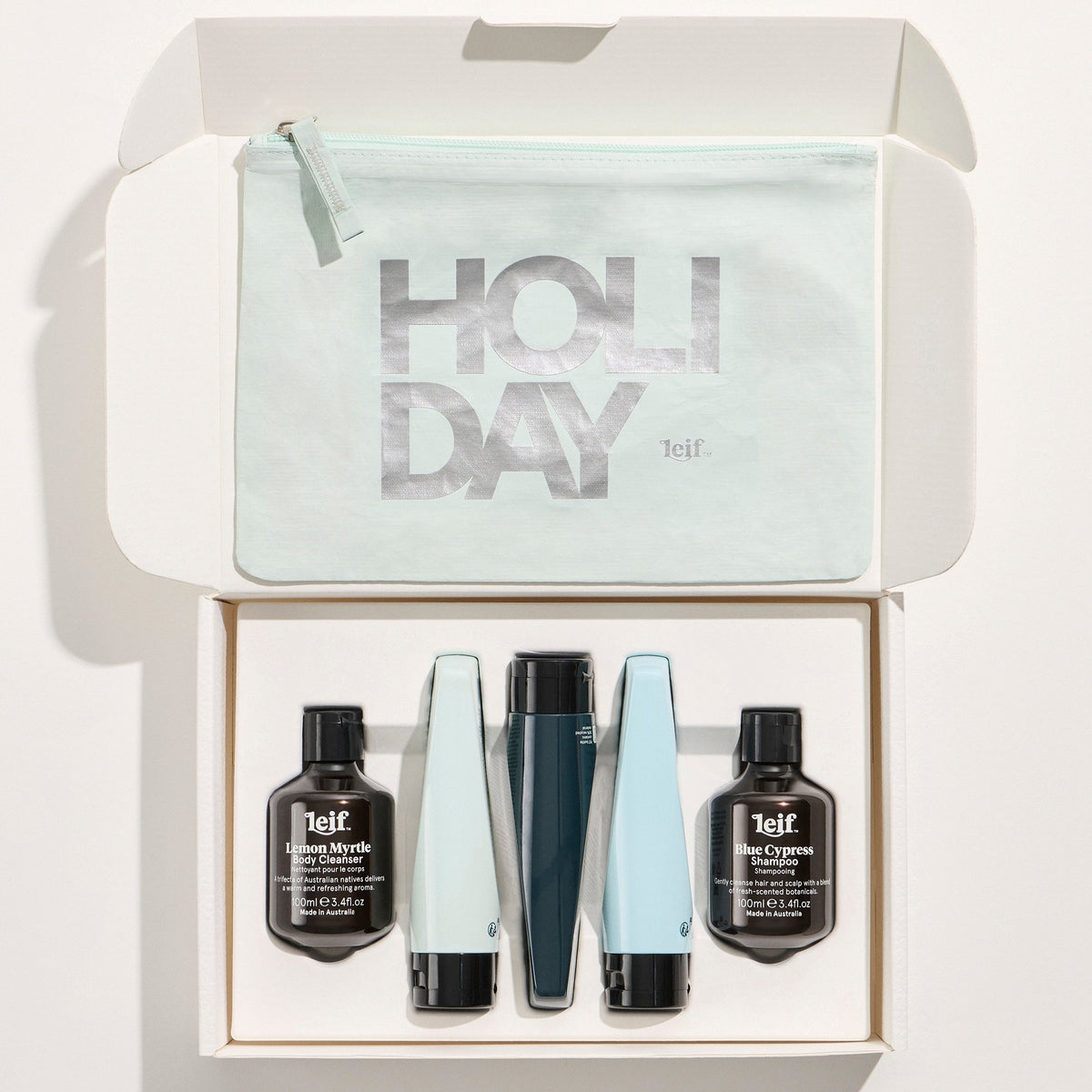 A boxed haircare gift set opened from above on a white background. The set includes a pale mint pouch printed with large metallic 'HOLIDAY' lettering, four bottles and tubes of hair product in black, dark teal, and light blue, and two small black bottles labeled telf.