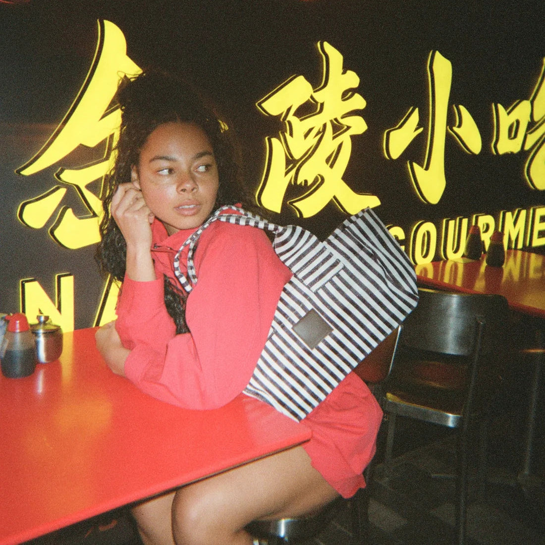 A woman sitting at a red table in a restaurant, wearing a coral pink dress and holding a black and white striped tote bag. The background features a dark wall with yellow Chinese characters and a neon bun outline.