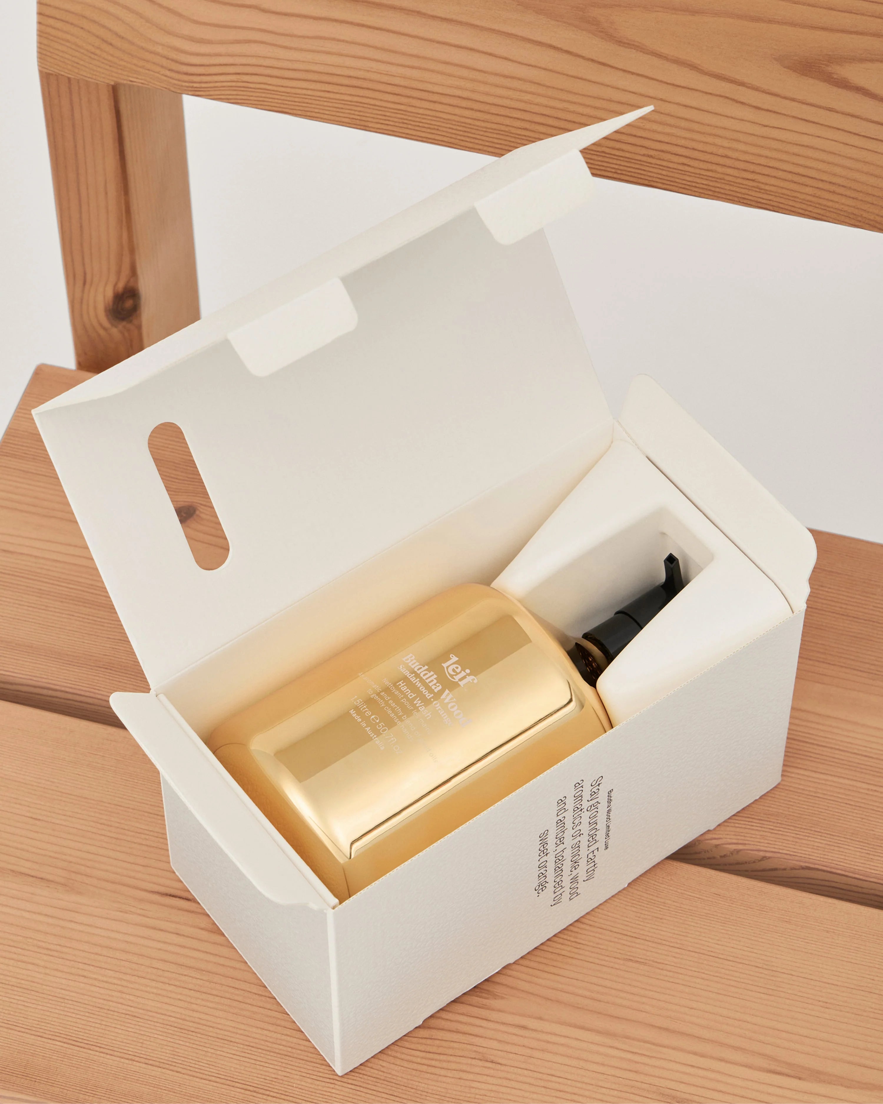 A gold cosmetic bottle with a black pump inside an open white gift box, placed on a light wood surface.