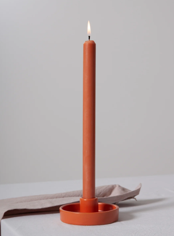 A tall, slender terracotta-colored candle standing upright in a matching round holder, with a small flame lit at the top. The candle and holder are placed on a light surface with a soft neutral fabric behind it against a plain light gray background.