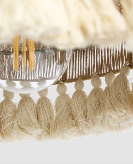 Close-up of a bohemian-style pendant light fixture featuring a fluffy cream faux-fur shade, a woven beige and brown fringe trim, and multiple cream tassels hanging below.