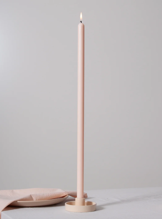 A tall, slender blush pink taper candle standing upright in a matching round holder on a light surface, with a folded napkin beside it against a soft neutral background.