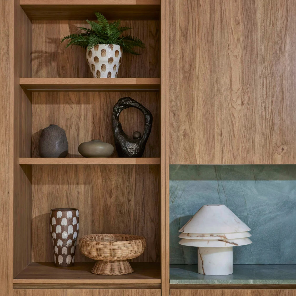 A styled interior scene showing a wooden shelving unit with decorative objects, including a potted fern, ceramic vases, a woven bowl, and a white tiered table lamp on a green stone surface.