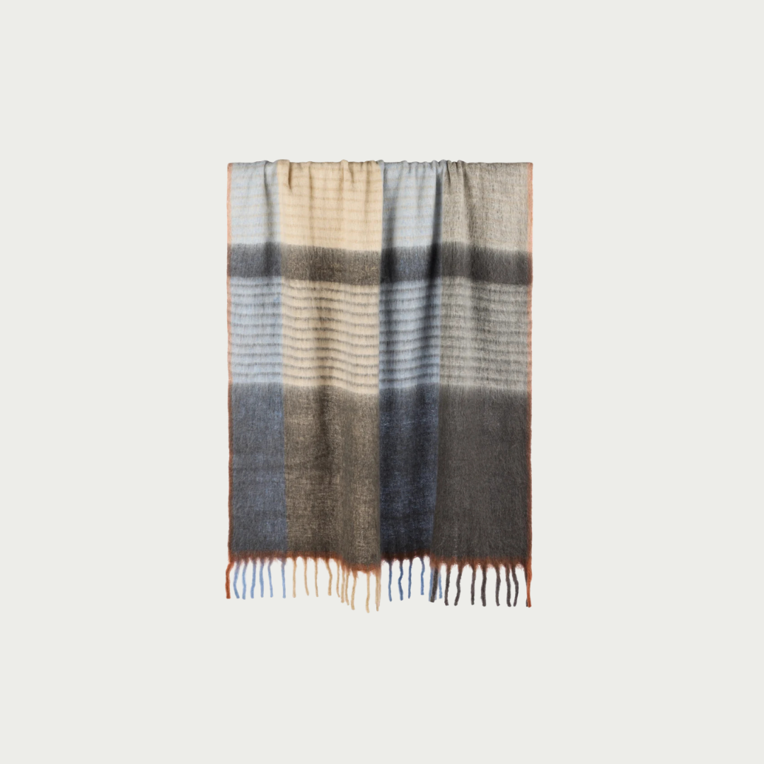 A striped knit scarf with a gradient design transitioning from beige and light gray at the top, through dark gray and blue-gray, to a darker charcoal at the bottom, finished with fringed ends.