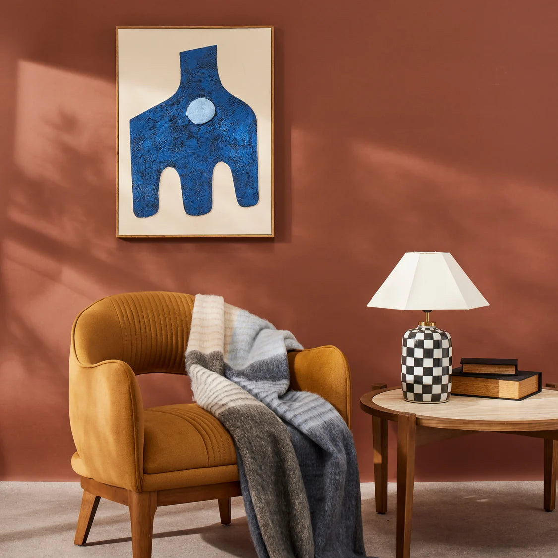 Brown armchair with a blanket, wooden side table with lamp and books, abstract blue artwork on brown wall.