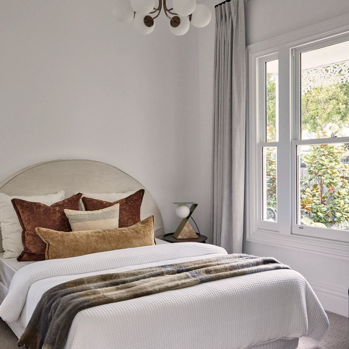 A styled bedroom interior featuring a bed with white textured bedding, brown and beige pillows, a gray faux-fur throw blanket, a light beige upholstered headboard, gray curtains, and a window looking out to greenery.